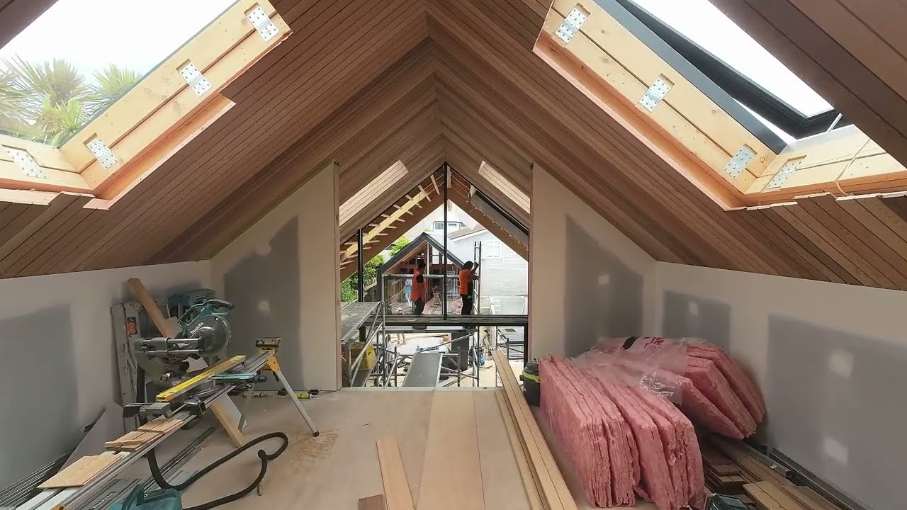 Drone view inside the upper floor of a house under construction: exposed timber ceiling lining, two skylights, a mitre saw and stacked insulation batts, looking out through the gable window to builders on the deck.